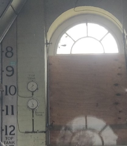 Interior of brick building illuminated by high window. There are gauges and numbers painted on the wall.