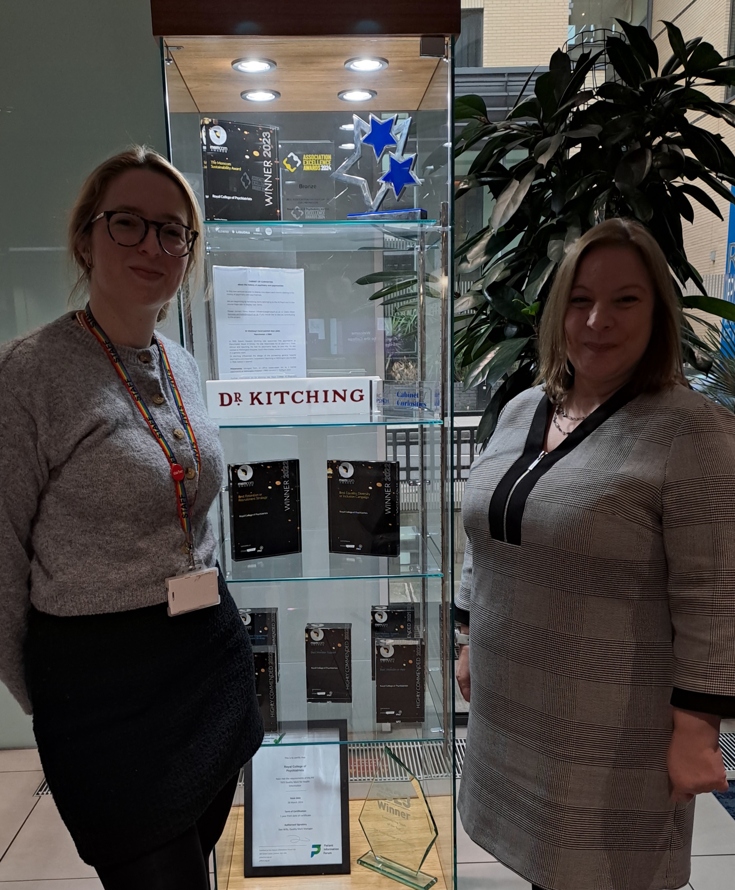 Photograph of two women standing either side of a tall glass cabinet containing various modern awards and one shelf with a sign reading Dr Kitching on it.
