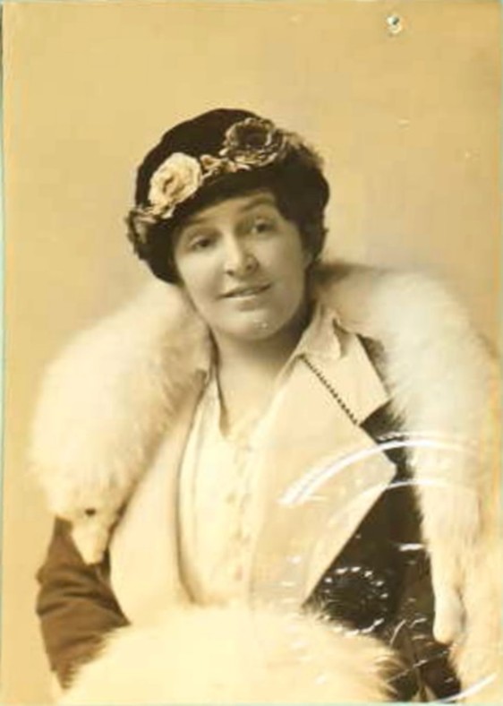 Sepia photo of a white woman in a suit with a white fox fur, fur muff and hat with flowers. She's smiling. 