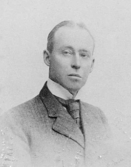 Balck and white faded photo of a white man in a suit with slicked back hair and a serious or arrogant expression.
