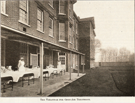 Black and white photo graph of a three story brick building with an awning over a paved area outside where patients lie in hospital beds attended by nurses.